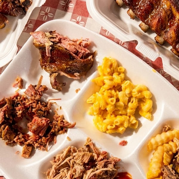 Barbecue platter with ribs, pulled pork, brisket, mac and cheese, and loaded fries on a red checkered tablecloth.