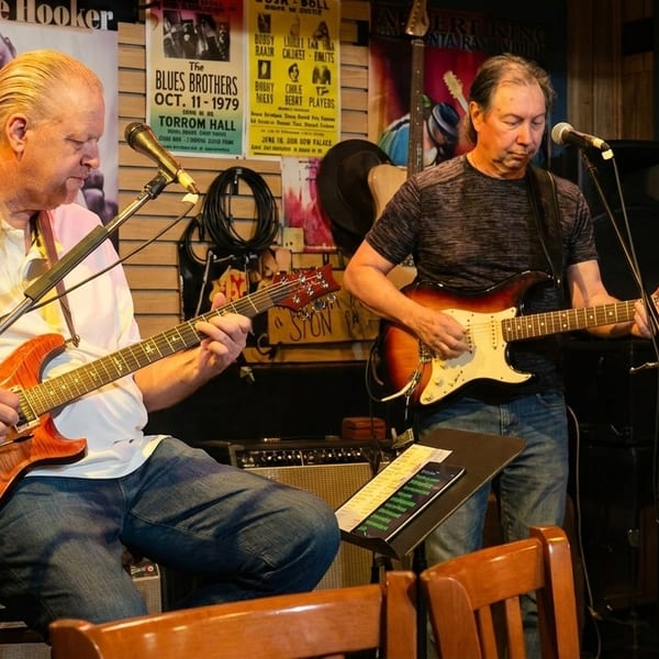 Two men playing electric guitars on stage with music posters in the background.